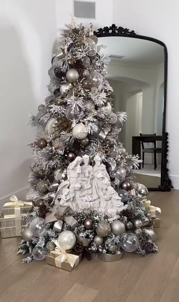 Silver and white Christmas tree decorated with metallic ornaments and a large white nativity scene nestled into the lower branches above wrapped gifts.