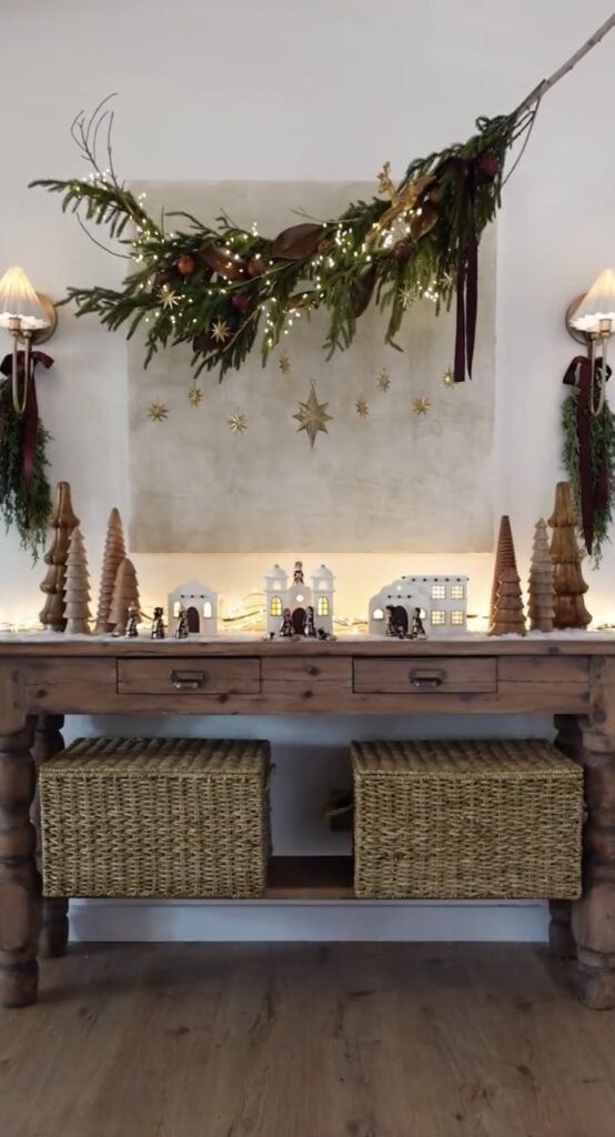 White Christmas village and nativity scenes arranged across a rustic wooden console with carved trees, warm lights, hanging stars, and greenery.