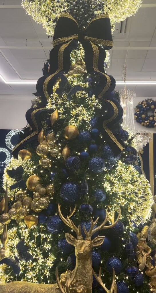 Glamorous Christmas tree decorated with navy blue and gold ornaments, metallic accents, warm lights, and a large statement bow.