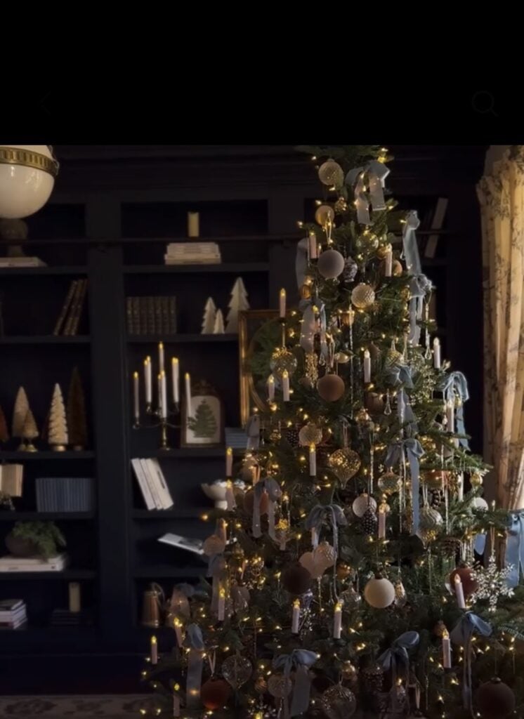 Traditional Christmas tree styled with dusty blue velvet bows, warm candle lights, vintage gold and brown ornaments, glass icicles, and soft golden lighting in a dark, book-lined living room.