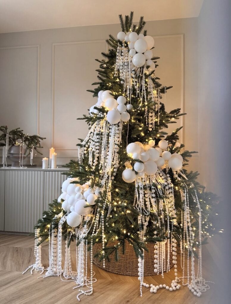 Traditional green Christmas tree decorated entirely in white with pale ornaments, ribbon, floral accents, and warm white lights.