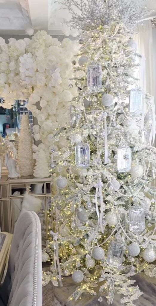 All-white Christmas tree topped with an oversized white satin bow and decorated with silver ornaments, frosted branches, and elegant hanging icicles.