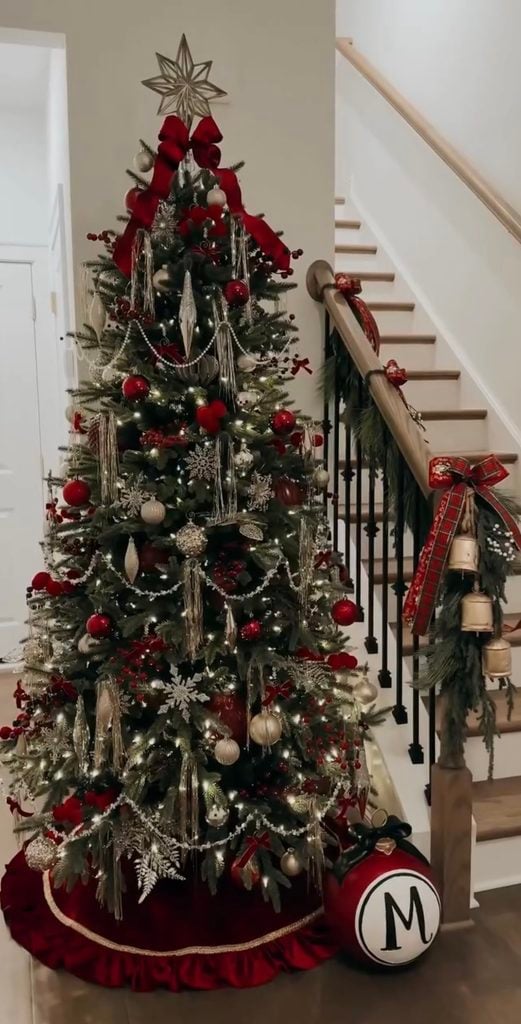 Christmas tree beside a staircase decorated with burgundy ribbon, red ornaments, silver accents, garland and a star topper.