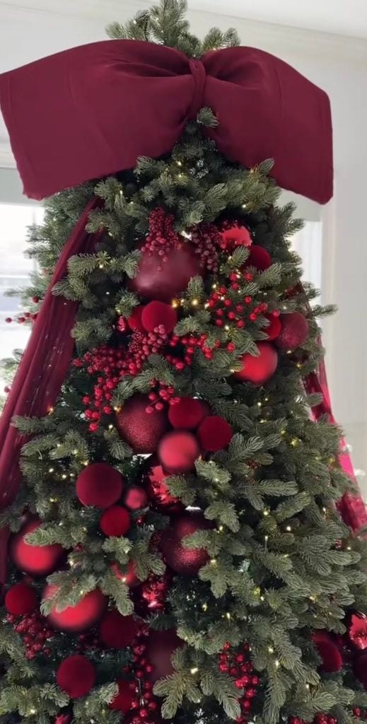 Burgundy Christmas tree topped with an oversized velvet bow and decorated with red ornaments, berries and warm lights.