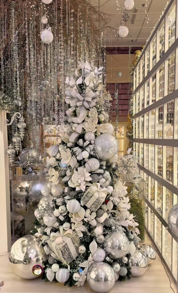 Glamorous green Christmas tree densely decorated with white flowers, oversized silver ornaments, white ribbon, glittering branches, and reflective metallic baubles.