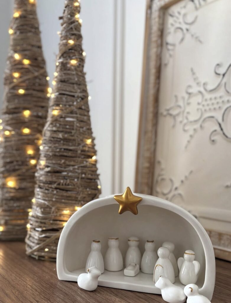 Small white ceramic nativity set with gold accents displayed beside lit wicker Christmas trees and framed neutral artwork.