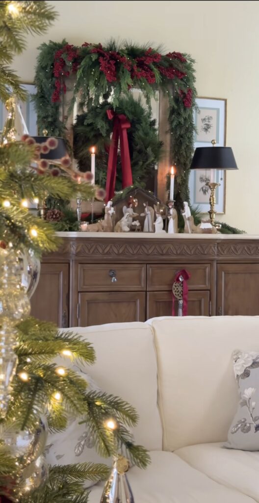Nativity set displayed on a wooden sideboard beneath a large evergreen wreath with red berries, burgundy ribbon, taper candles, and a decorated Christmas tree nearby.