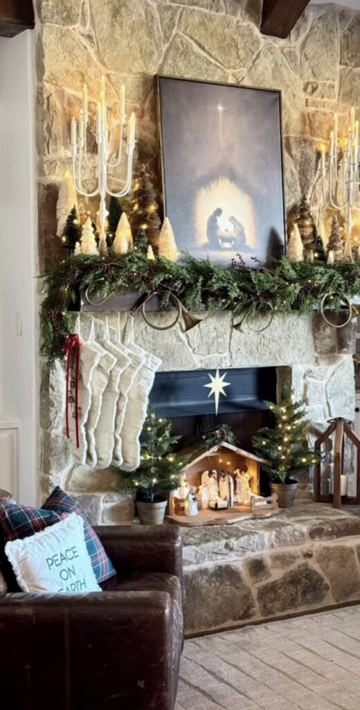 Stone fireplace decorated with a nativity scene, evergreen garland, bottle-brush trees, glowing candles, white stockings, and a framed Holy Family print above the mantel.
