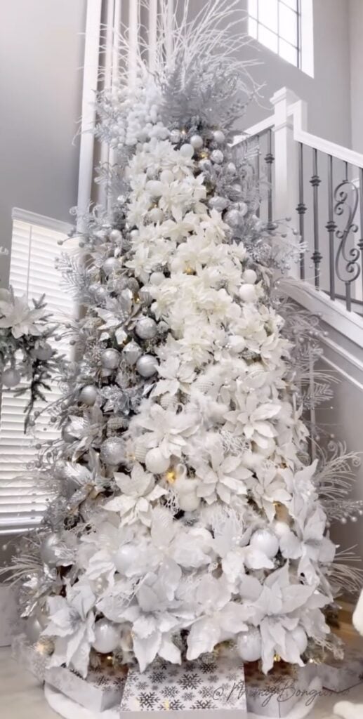 Tall Christmas tree layered with white poinsettias, silver ornaments, frosted branches, snowy foliage, and warm white lights beside a staircase.