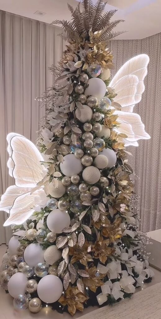 Whimsical Christmas tree decorated with oversized white fairy wings, pearl and silver ornaments, gold leaves, metallic foliage, and large white baubles.