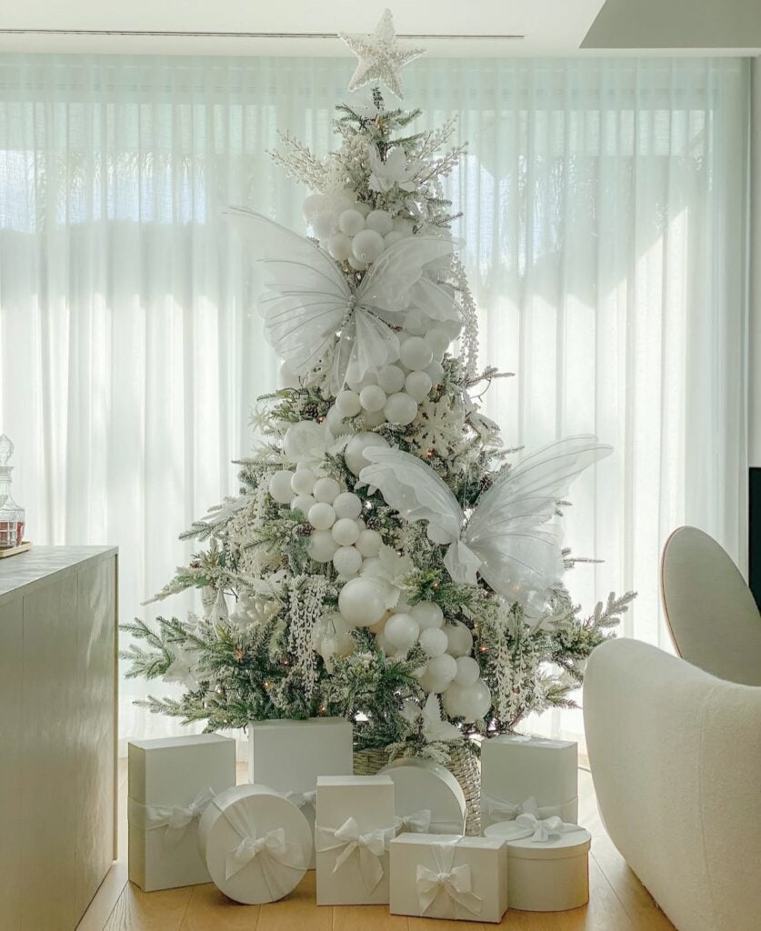 Flocked Christmas tree decorated with oversized white fairy wings, clusters of white ornaments, snowy foliage, a glittering star topper, and white gifts.