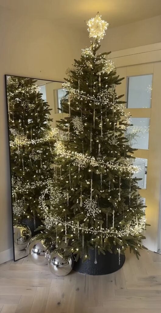 Simple green Christmas tree decorated with delicate white light garlands, hanging crystal icicles, a glowing star topper, and oversized silver ornaments at the base.