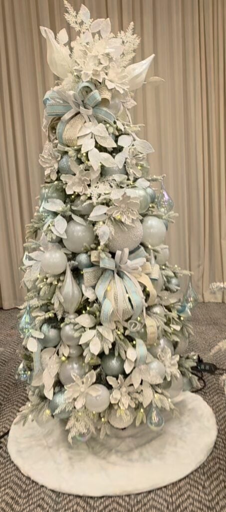 Flocked Christmas tree decorated with oversized light blue and white ornaments, pale blue ribbon, white poinsettias, frosted greenery, and warm white lights on a white faux-fur tree skirt.