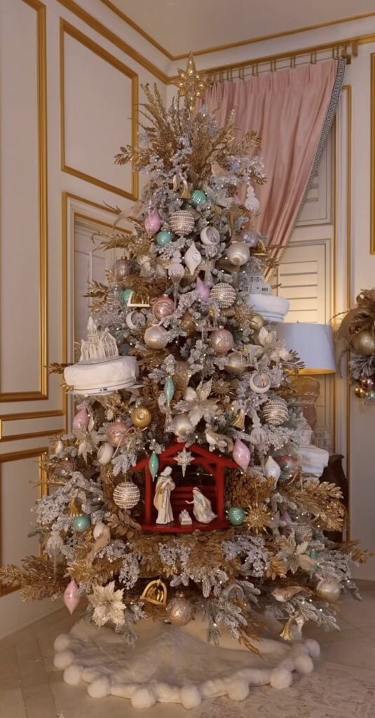 Pastel Christmas tree decorated with gold, pink, and turquoise ornaments with a red manger scene and nativity figures built into the lower branches.