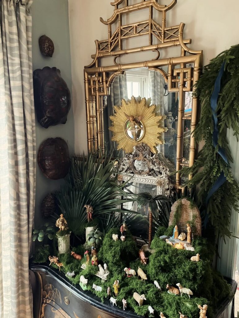 Elaborate miniature nativity scene spread across moss-covered hills beneath a gold pagoda mirror with palm fronds, animals, shepherds, and evergreen garland.
