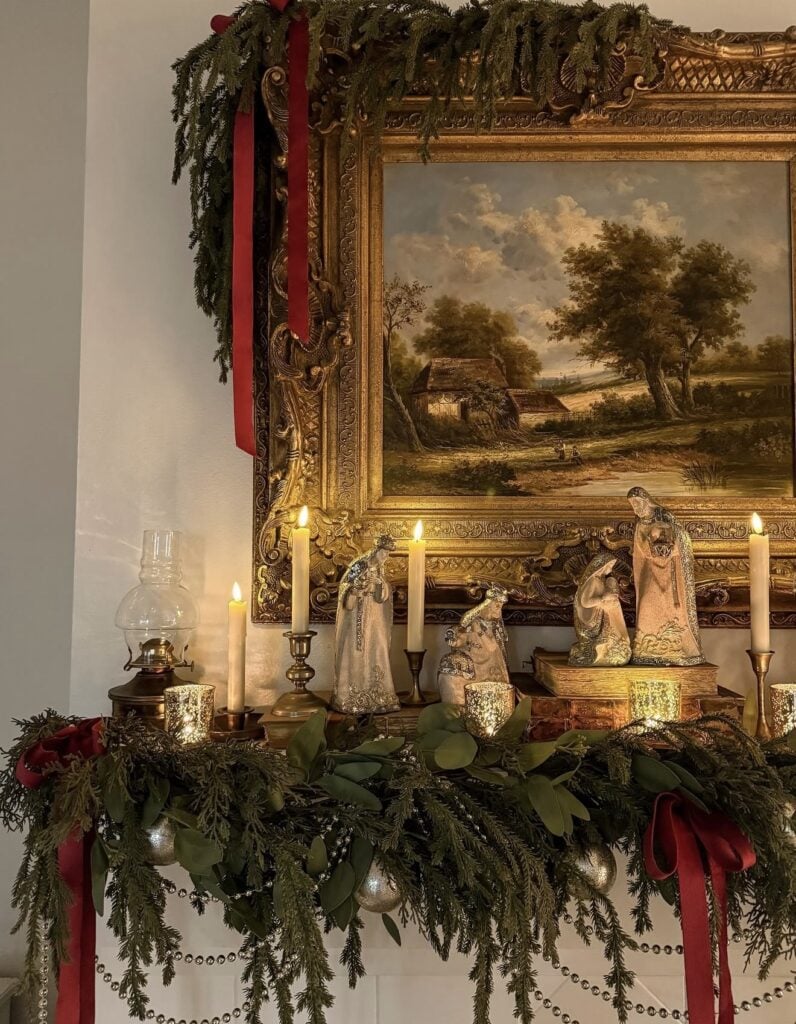 Traditional nativity figures displayed on a candlelit Christmas mantel with layered evergreen garland, gold ornaments, red velvet ribbon, and an antique landscape painting.