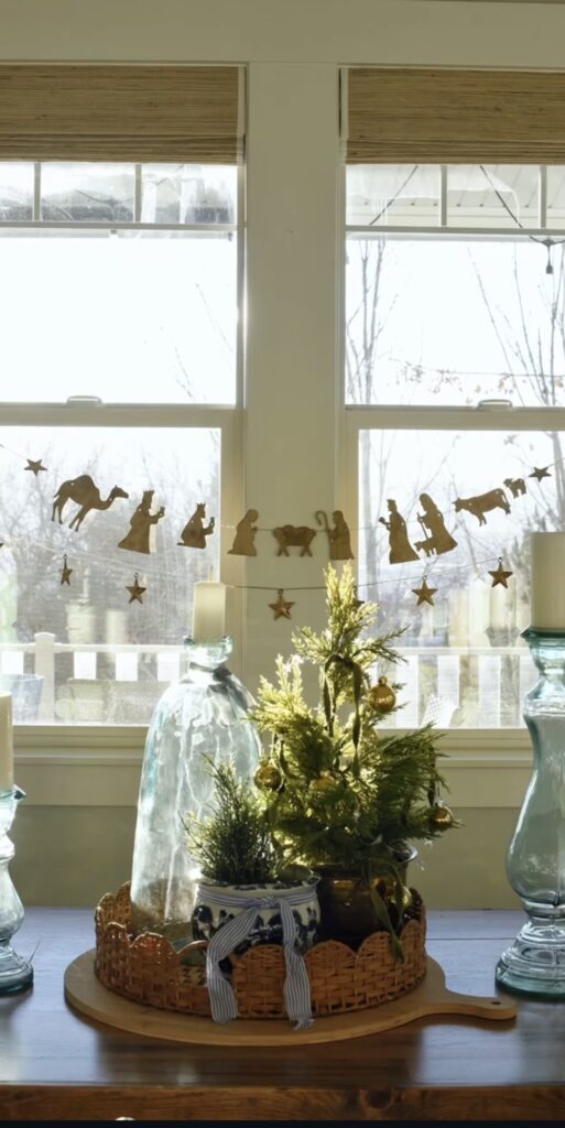 Nativity silhouette garland with stars hung across bright windows above a wooden table styled with miniature evergreen trees, glass candleholders, and a woven tray.