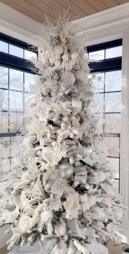 Elegant all-white Christmas tree decorated with layered white ornaments, snowy branches, ribbon, florals, reflective details, and warm white lights.