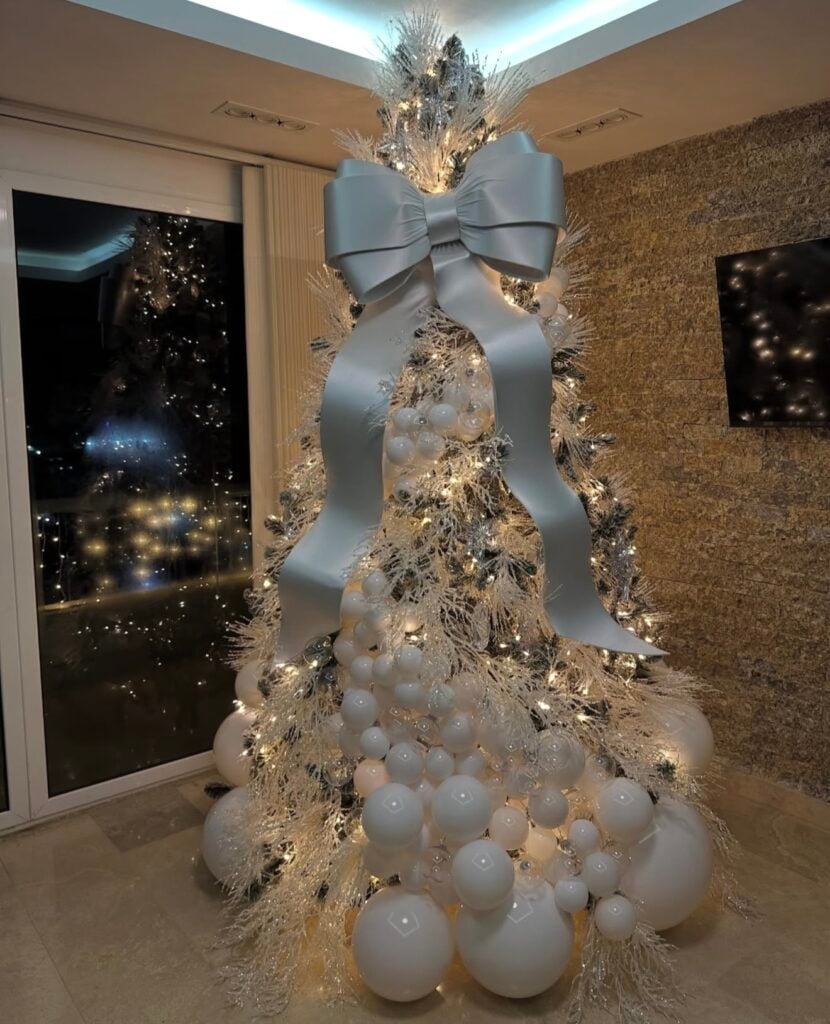 White artificial Christmas tree covered in clusters of glossy white ball ornaments, feathery white branches, warm lights, and an oversized silver-white satin bow.