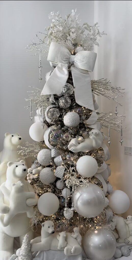 White Christmas tree decorated with layered white ornaments, snowy accents, textured ribbon, reflective details, and glowing holiday lights.