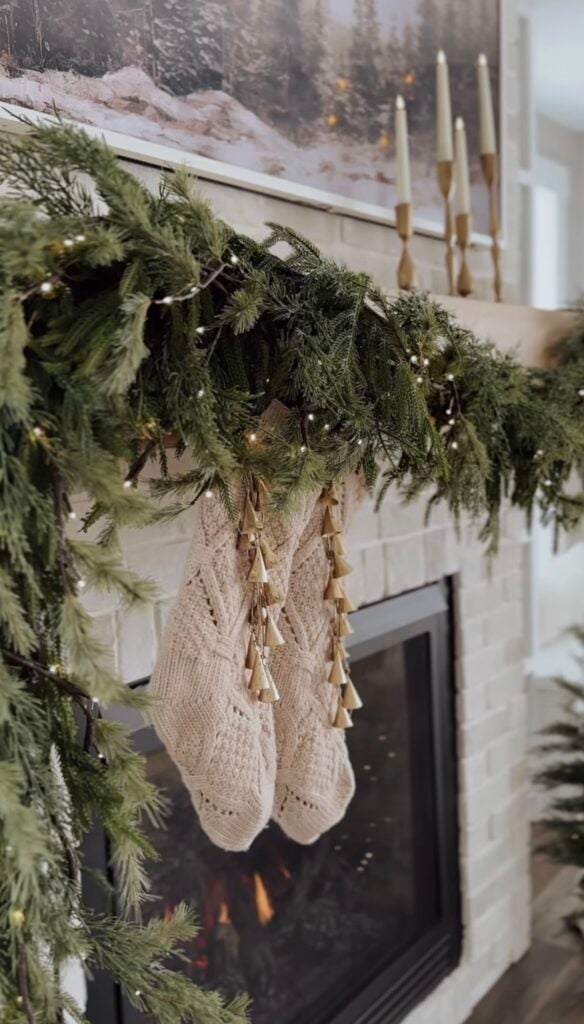 Christmas mantel with cream knit stockings, bells, greenery, and soft traditional holiday styling.