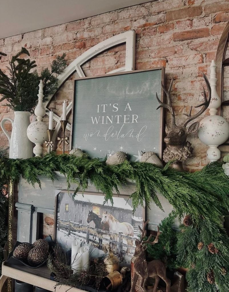 Layered Christmas fireplace mantel decorated with lush greenery, candles, ornaments, and holiday accents at different heights.