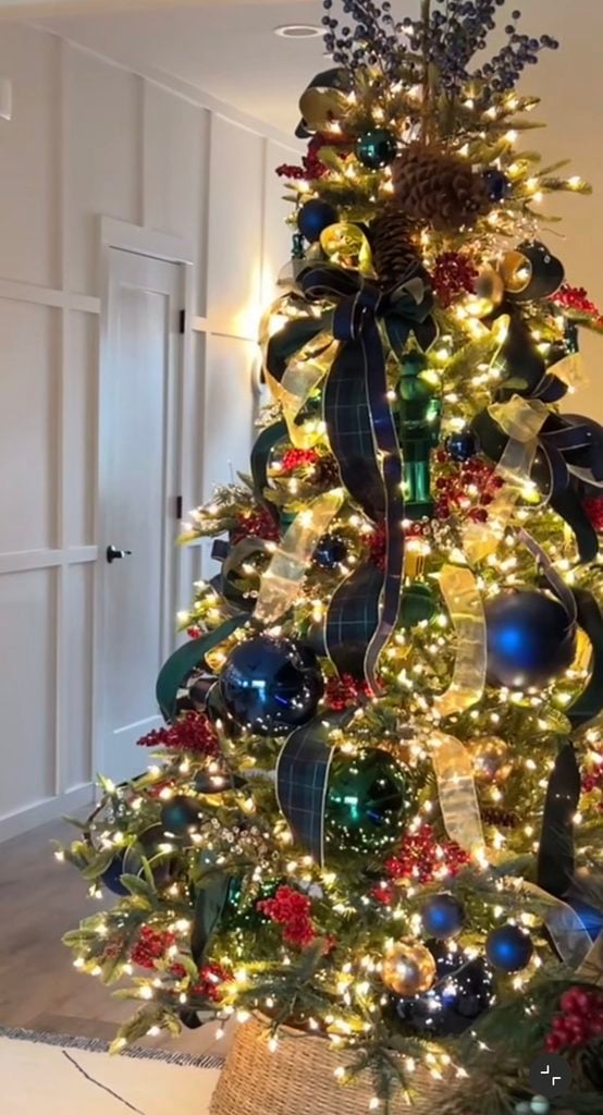 Ralph Lauren Christmas tree with navy, hunter green, burgundy, and gold ornaments, plaid ribbon, red berries, pine cones, warm lights, and a woven tree basket.
