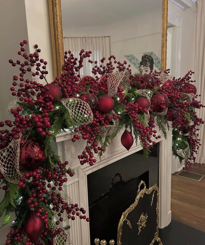 Luxe red Christmas fireplace mantel decorated with rich greenery, dramatic red accents, ribbon, and elegant holiday details.