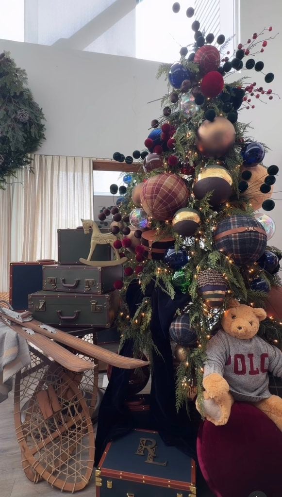 Ralph Lauren Christmas tree decorated with oversized plaid ornaments, navy and burgundy accents, velvet ribbons, leather details, a Polo teddy bear, vintage luggage, and snowshoes.
