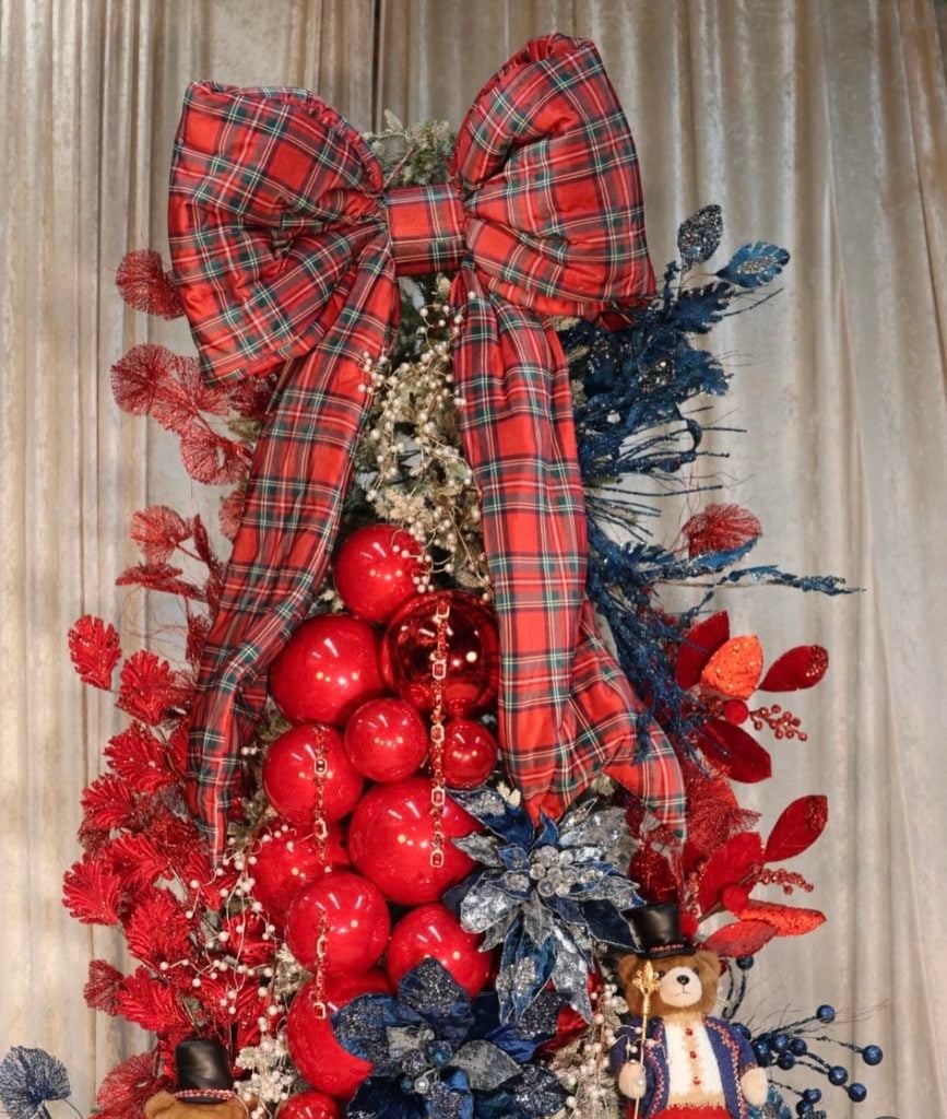 Ralph Lauren-inspired Christmas tree topped with an oversized red tartan bow and decorated with glossy red ornaments, navy accents, metallic florals, and a teddy bear nutcracker.
