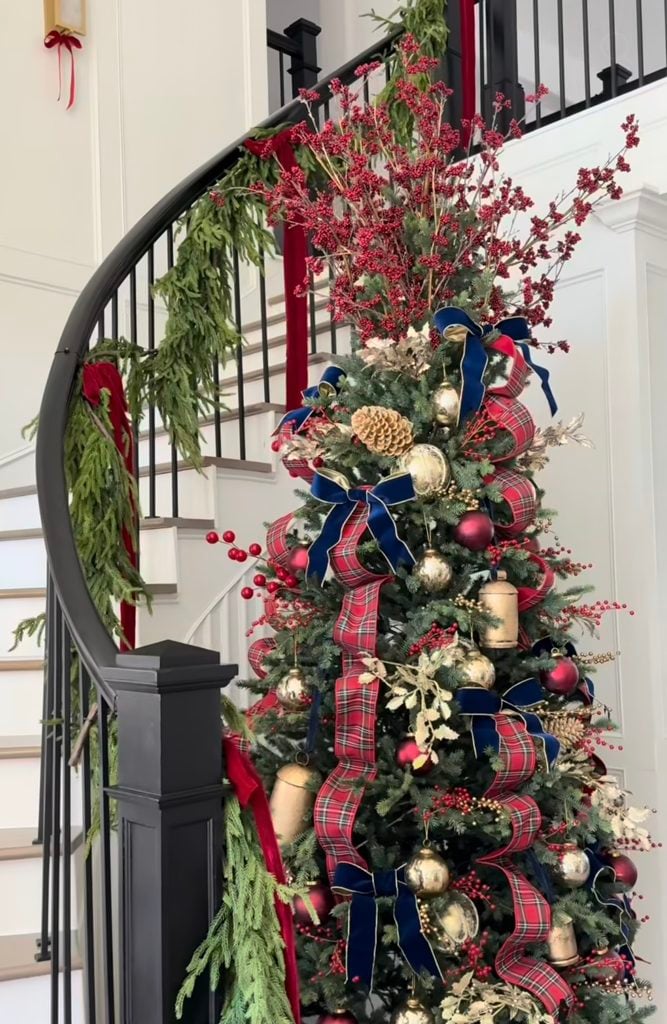 Ralph Lauren Christmas tree beside a staircase decorated with red tartan ribbon, navy velvet bows, red berries, brass ornaments, pine cones, and fresh greenery.
