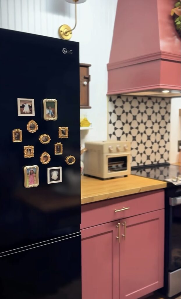 Black refrigerator with a small fridge gallery wall of miniature gold and white framed family photos in a colorful pink kitchen.