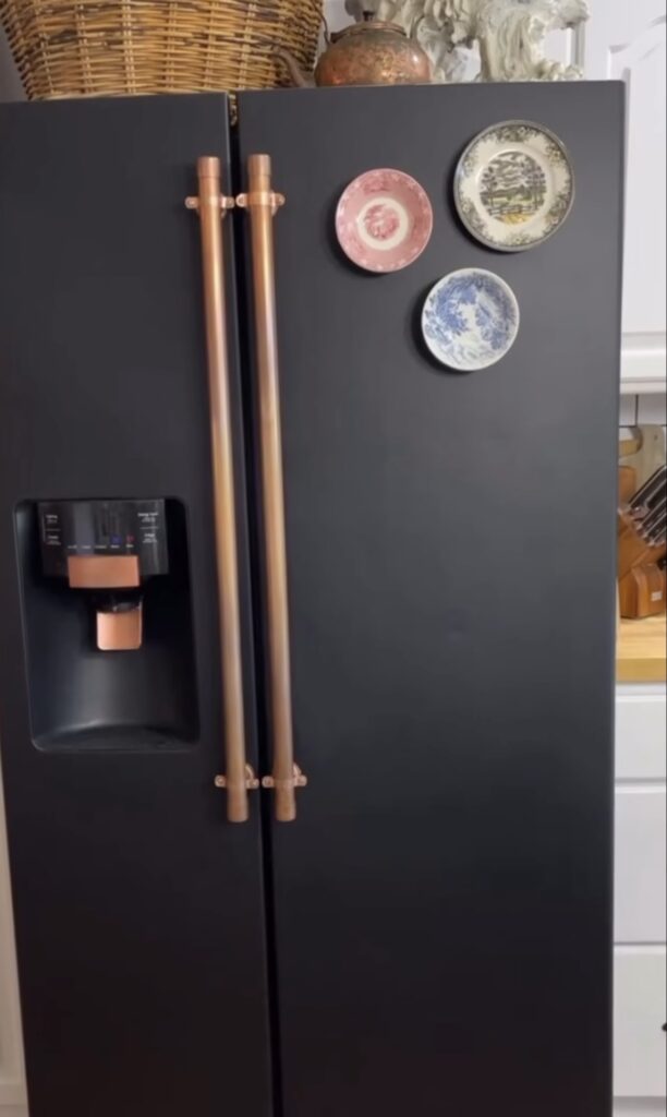 Black refrigerator with copper handles and a simple gallery wall of three decorative vintage plates in pink, blue and cream.