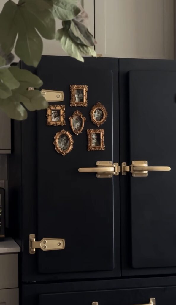 Black vintage-style refrigerator with brass hardware and a small gallery wall of ornate gold-framed family photos.