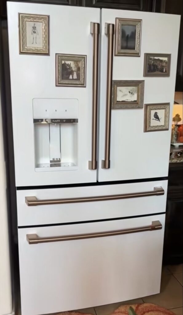 Minimal fridge gallery wall on a white refrigerator with small vintage-style framed artwork arranged around bronze refrigerator handles and a water dispenser.