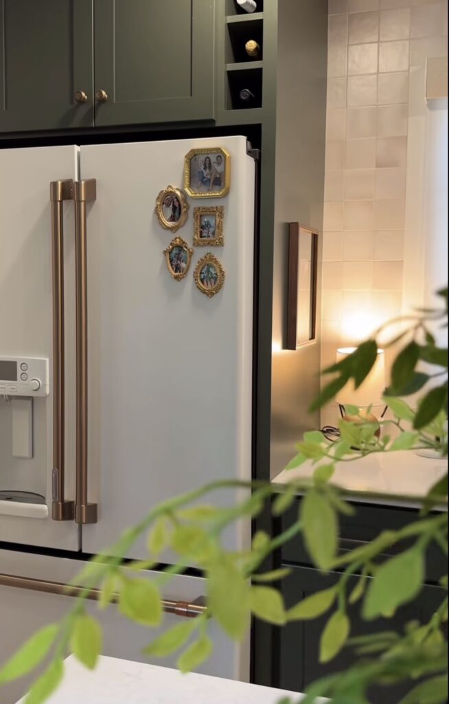 White refrigerator with a small fridge gallery wall of ornate gold family photo frames in a dark green and white kitchen.