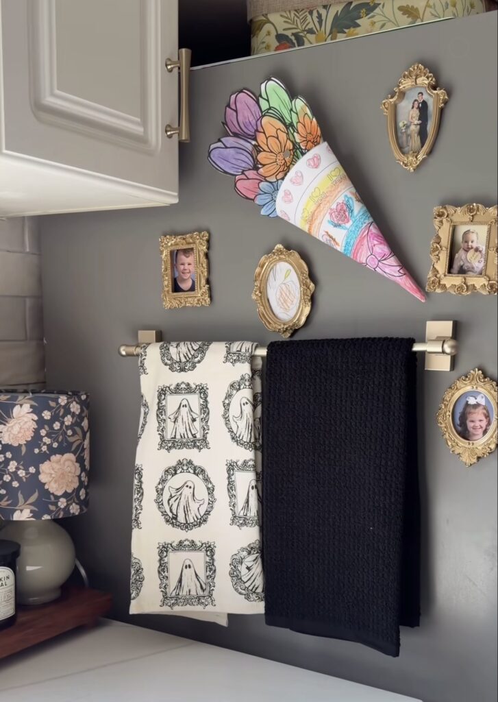 Side of a gray refrigerator decorated with gold-framed family photos, children's artwork and a magnetic towel bar with Halloween hand towels.