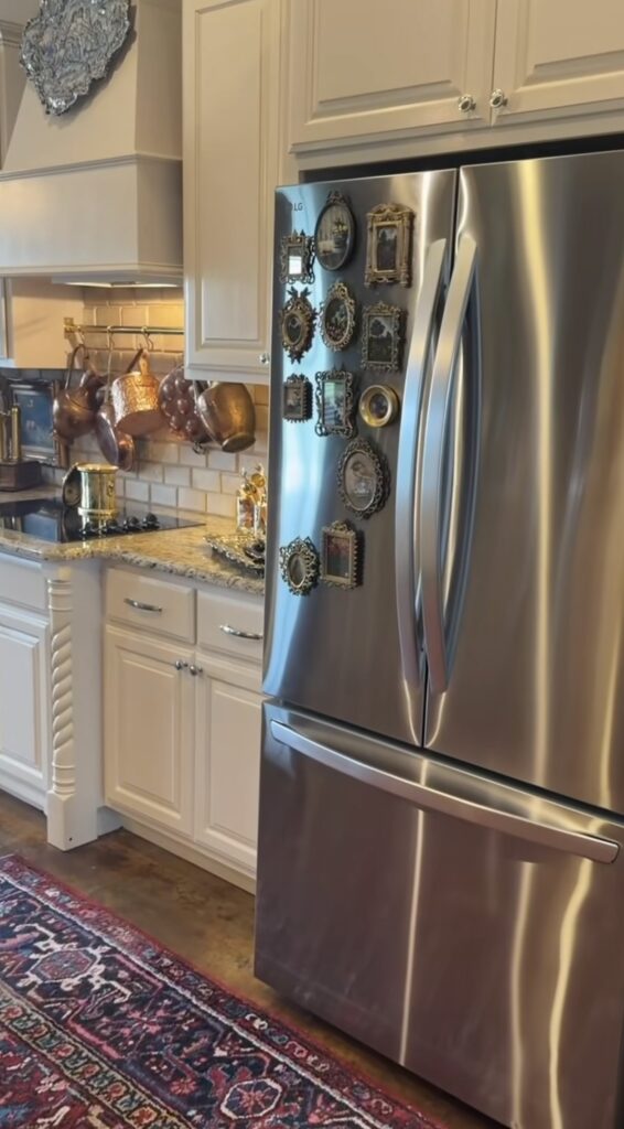 Stainless steel refrigerator with a gallery wall of ornate vintage gold frames displayed on the side beside a traditional kitchen with copper cookware.