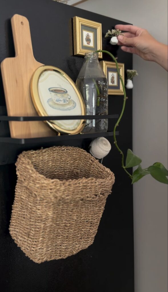 Creative fridge gallery wall on a black refrigerator with tiny gold frames, vintage artwork, a cutting board, glass bottle, greenery and woven storage basket.