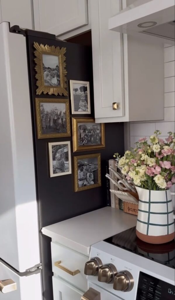 Family photo gallery wall displayed on the black side of a refrigerator with mismatched gold frames beside white kitchen cabinets and fresh flowers.