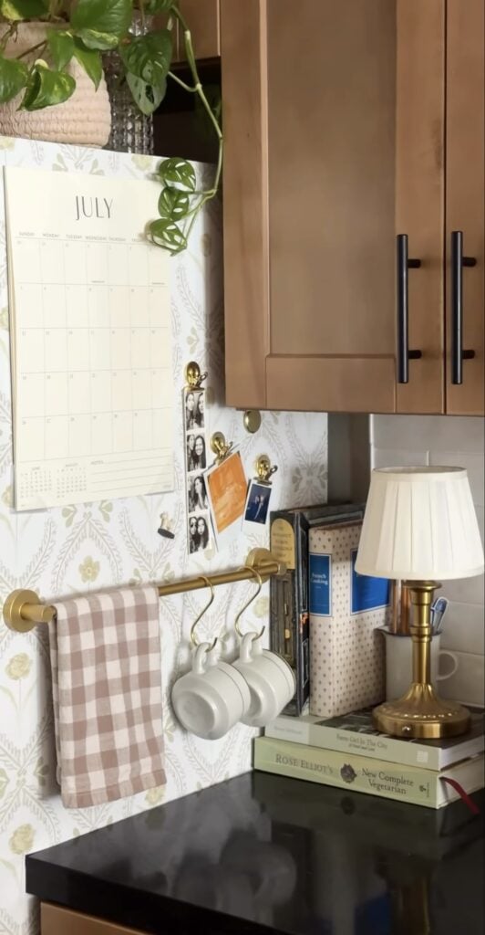 Wallpapered side of a refrigerator with a brass towel bar, hanging mugs, photographs and a calendar.