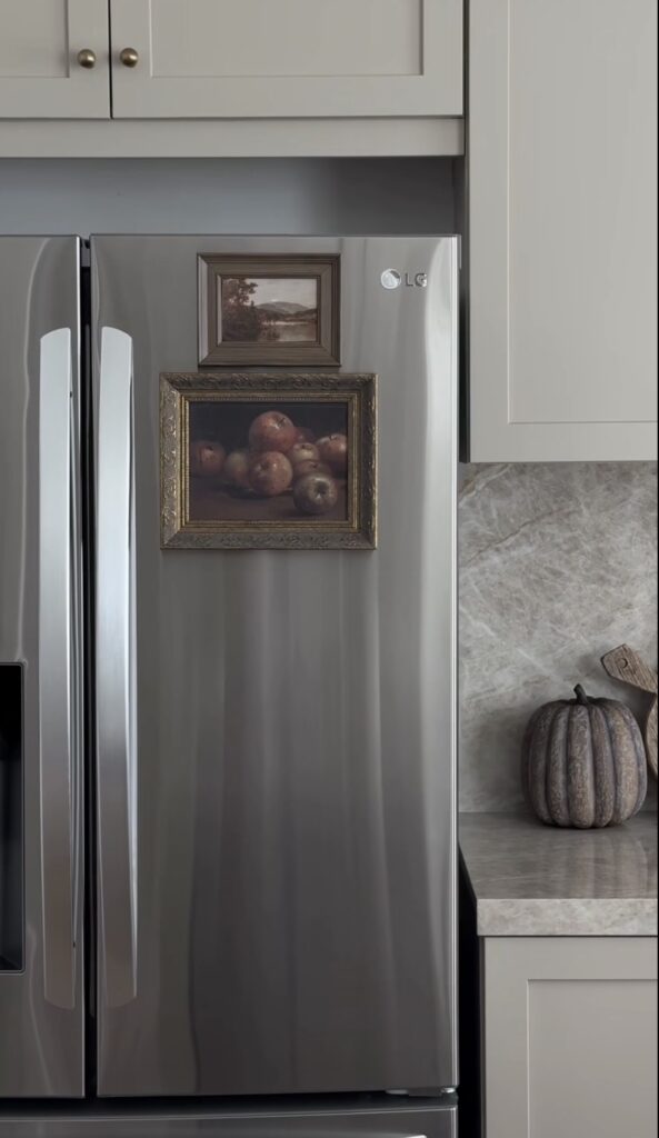 Minimal fridge gallery wall on a stainless steel refrigerator with two framed vintage-style landscape and still life paintings in a neutral kitchen.