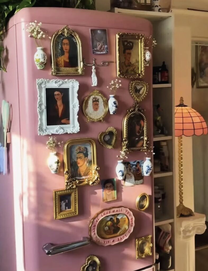 Maximalist fridge gallery wall on a pink refrigerator filled with ornate gold frames, Frida Kahlo artwork, tiny decorative vases, flowers and eclectic vintage-inspired decor.