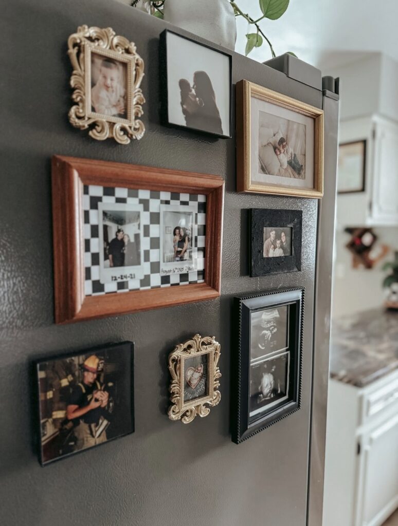 Family photo gallery wall on the side of a dark refrigerator with mismatched black, wood and ornate gold frames, including family photos and an ultrasound picture.