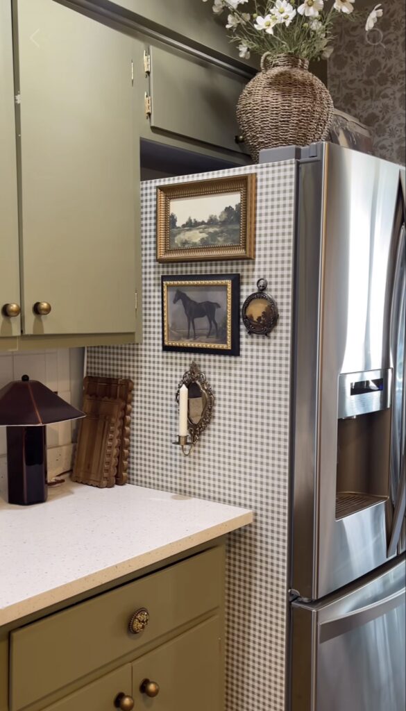 Side of a refrigerator covered in neutral gingham wallpaper and decorated with framed landscape art, horse artwork, an ornate mirror and a candle sconce.