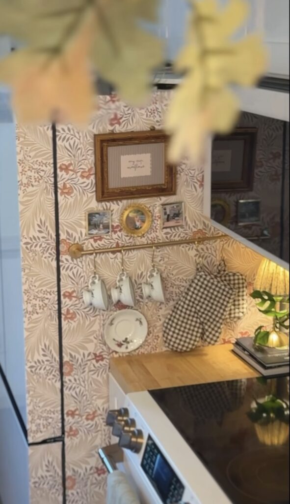 Wallpapered fridge gallery wall with floral wallpaper, small framed photos and art, a brass towel rod, hanging teacups and gingham oven mitts.