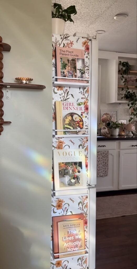 Side of a refrigerator decorated with floral motifs and white book racks holding cookbooks and magazines