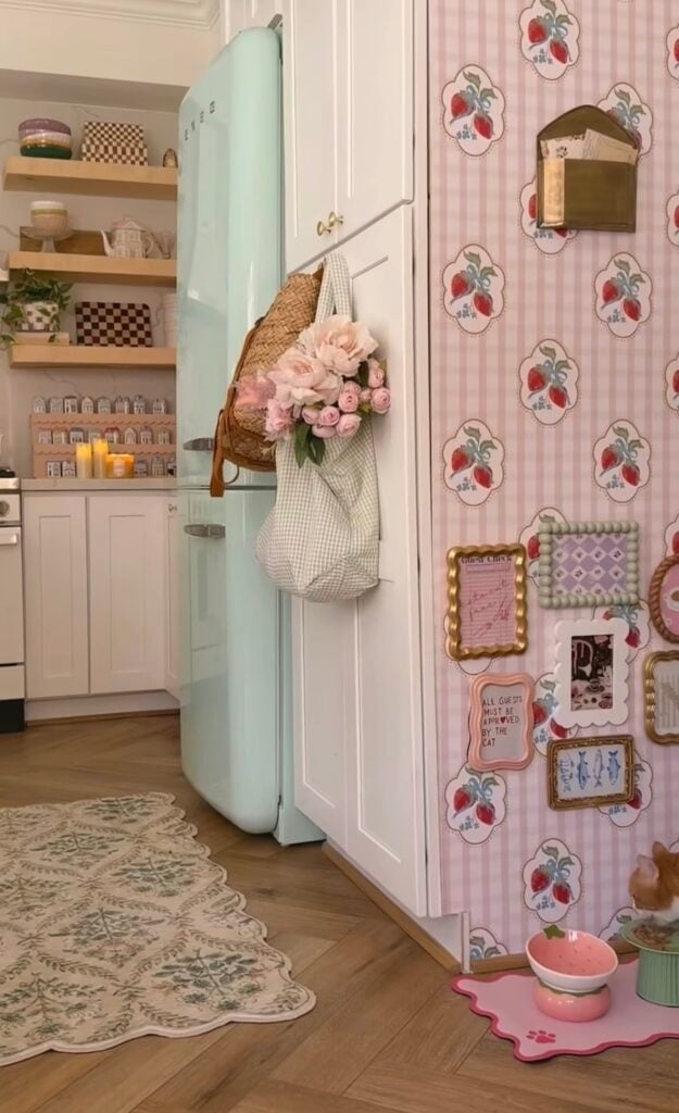 Pink cat feeding station gallery wall in a kitchen with strawberry wallpaper, framed cat art, decorative signs, pink food bowl, and a cat eating nearby.