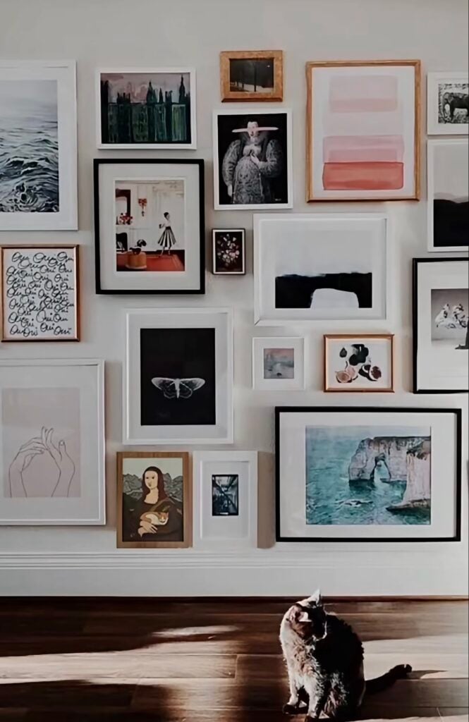 Large eclectic gallery wall with mixed framed artwork and a cat sitting on the wood floor below.
