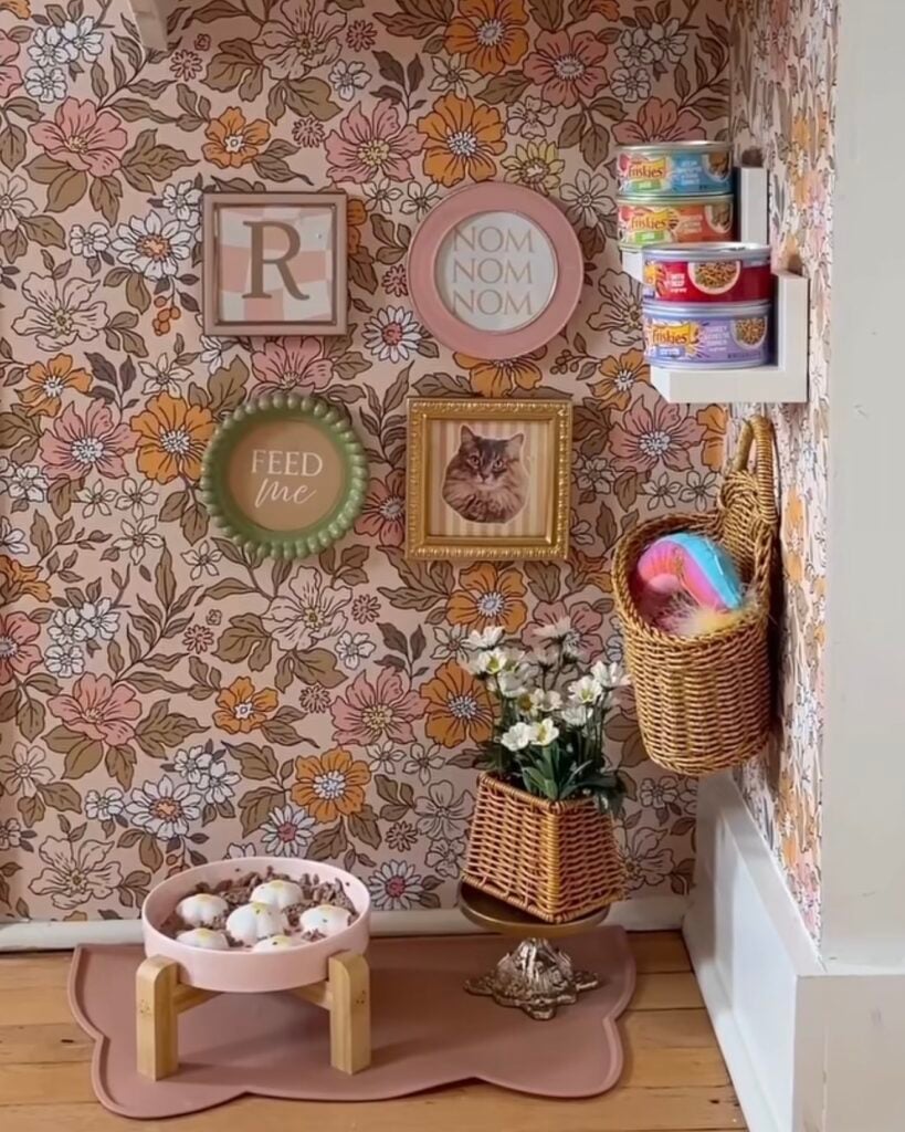 Cat feeding station gallery wall with vintage floral wallpaper, framed cat portrait, playful feeding signs, pink raised food bowl, and wall-mounted cat food storage.